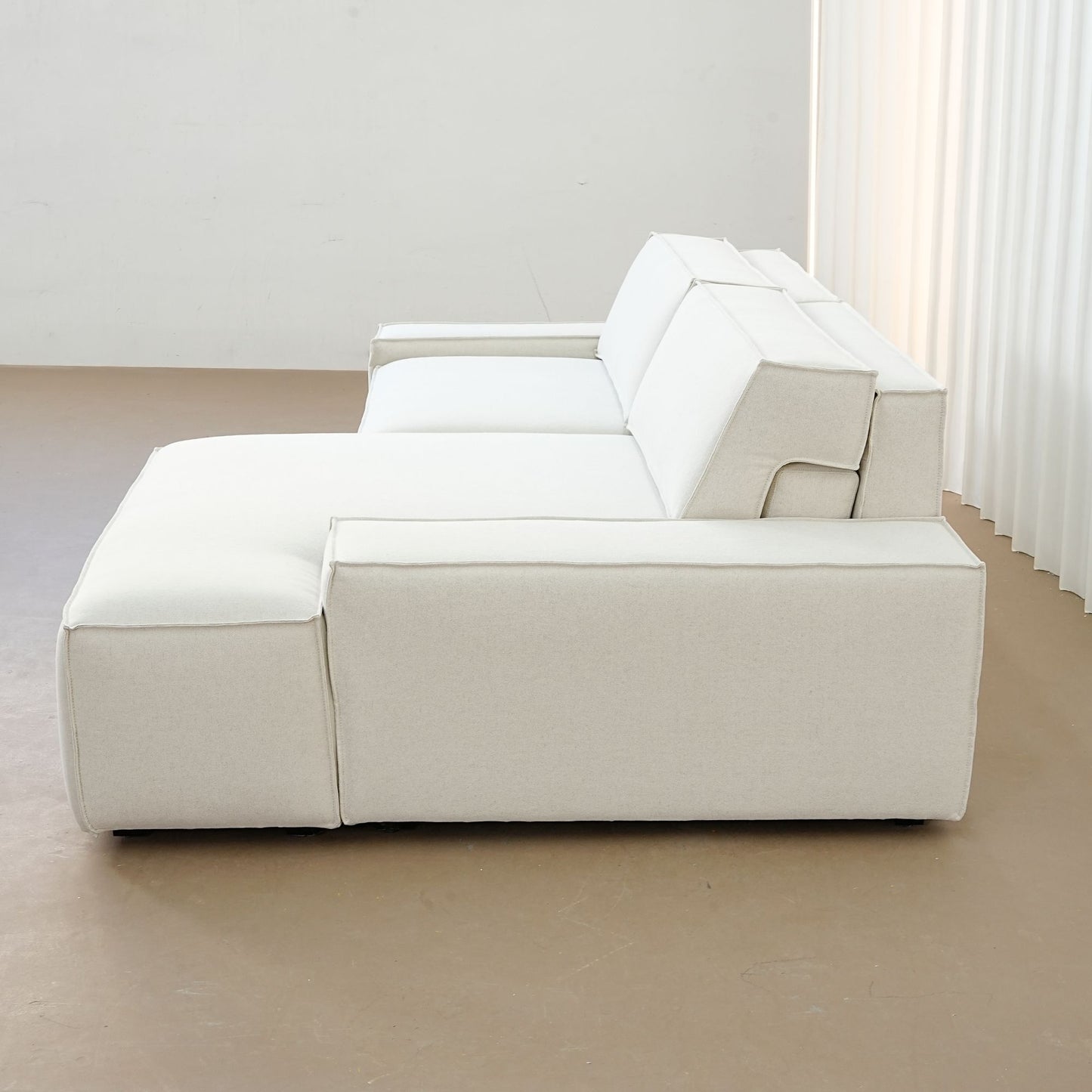 Beige sectional sofa in a minimalistic room with white walls and curtains.