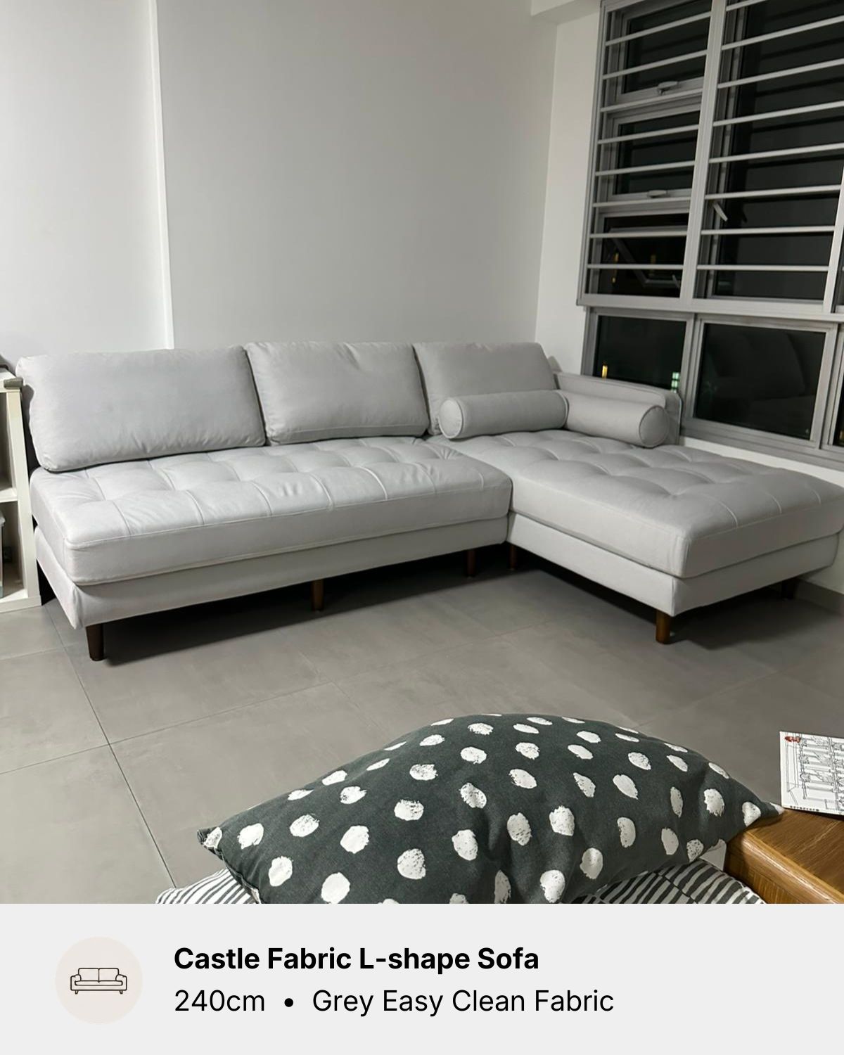 A light grey L-shaped fabric sofa in a modern living room setting, with a text overlay at the bottom identifying the model as Castle Fabric L-shape sofa and noting the fabric is grey and easy to clean. 