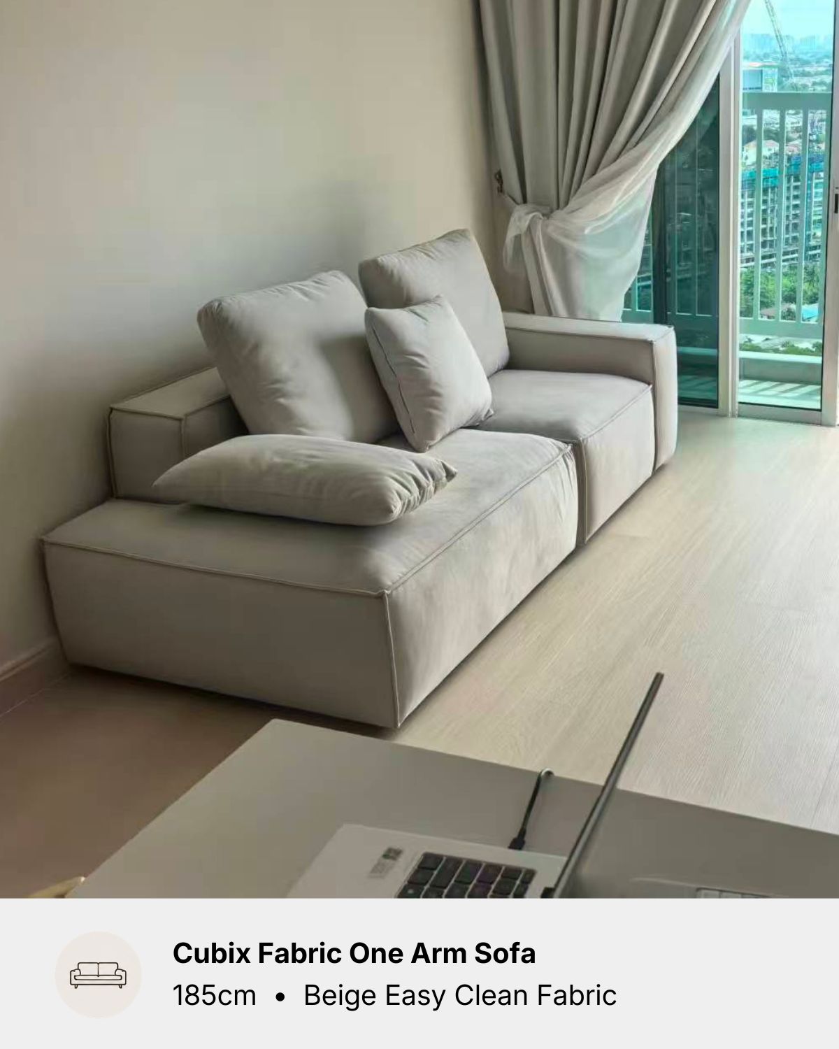 A light beige modular fabric one-arm sofa with matching throw pillows, shown in a modern living room setting with light wood floors and a window with curtains.