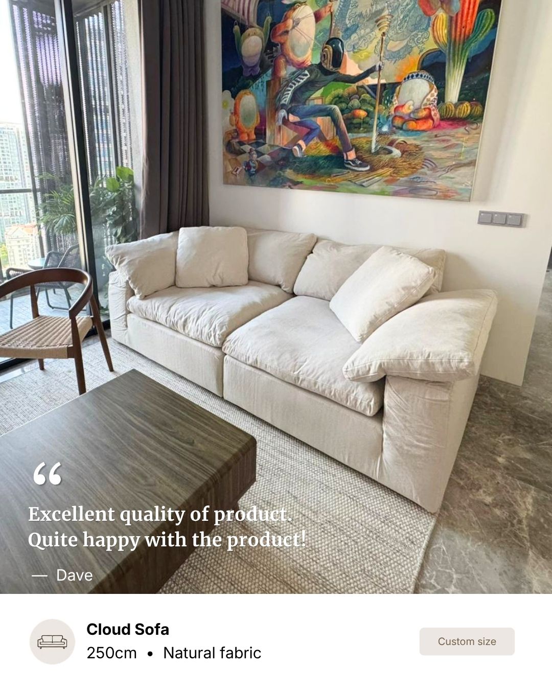 A beige colored Cloud Sofa with natural fabric cushions, shown in a modern living room setting with a dark wood coffee table, a woven rug, and a colorful painting on the wall.
