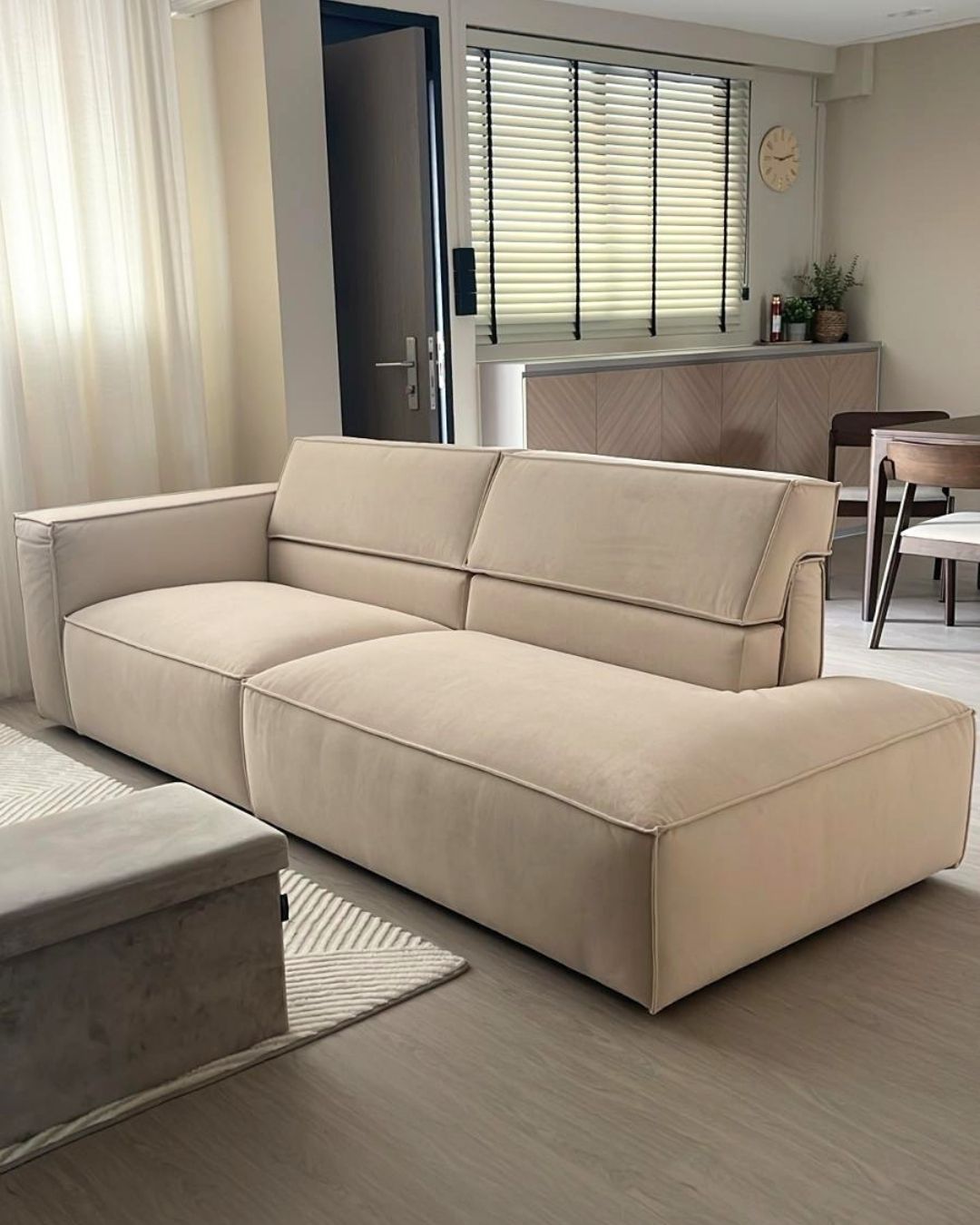 A modern beige high-back one arm sofa in a contemporary living room setting, featuring a low-profile design with wide armrests and a chaise lounge on the right side.