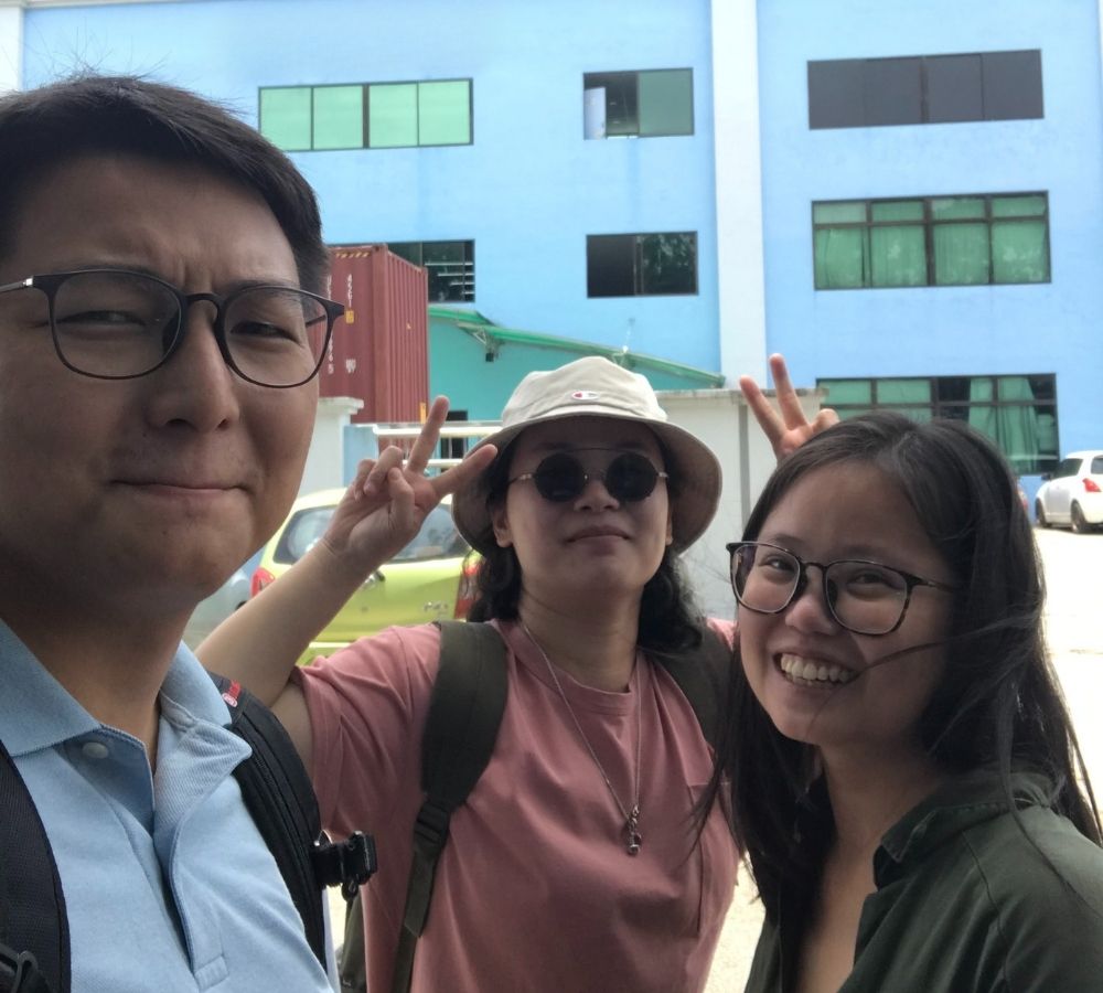 Three people taking a selfie outdoors in front of a light blue sofa factory building. The person on the left wears glasses and a light blue shirt, the center person wears a beige bucket hat, sunglasses, and a pink shirt, and the person on the right wears glasses and a dark green top.