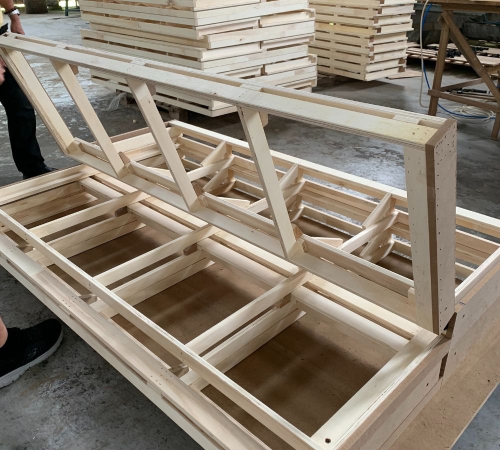 A partially assembled light natural wood frame or crib structure photographed in a workshop or warehouse setting, with stacks of similar wooden pieces and a concrete floor visible in the background. Parts of a person's legs and shoes are visible at the left edge.