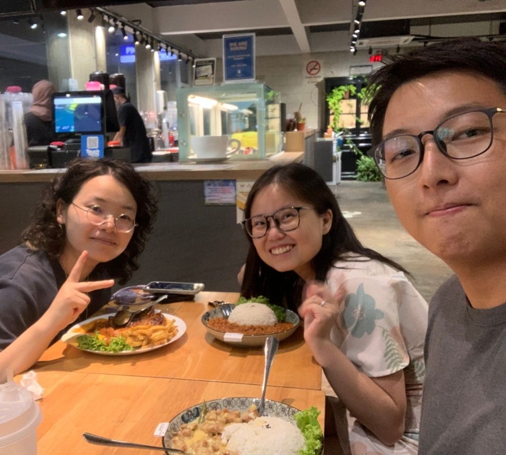 Three people taking a selfie at a restaurant table with food, including a plate of fried fish, a bowl of rice with meat, and another dish, in a modern indoor dining space.