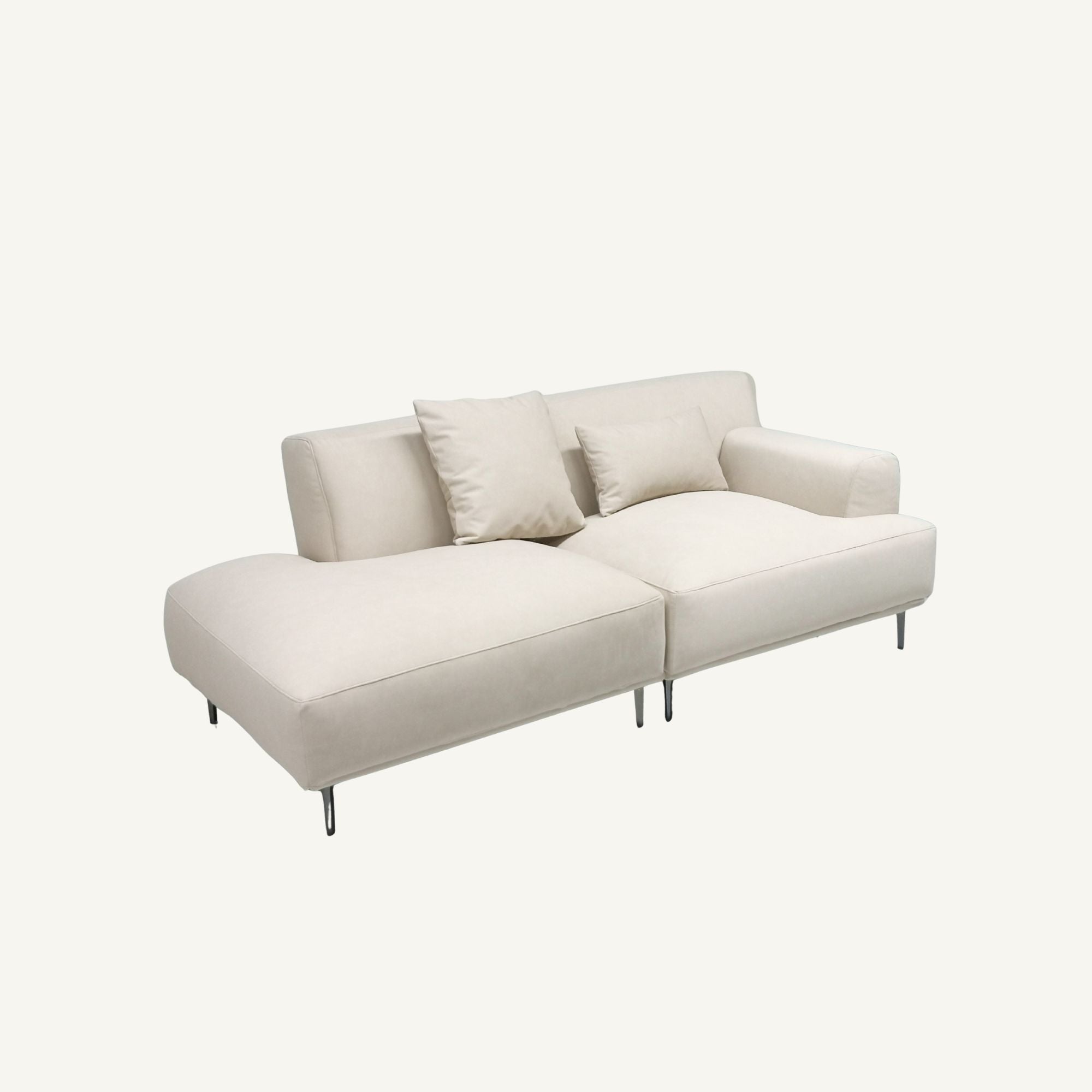 A modern modular sectional sofa in a light cream or off-white fabric, featuring two loose throw pillows and slim metal legs, shown against a plain off-white background.