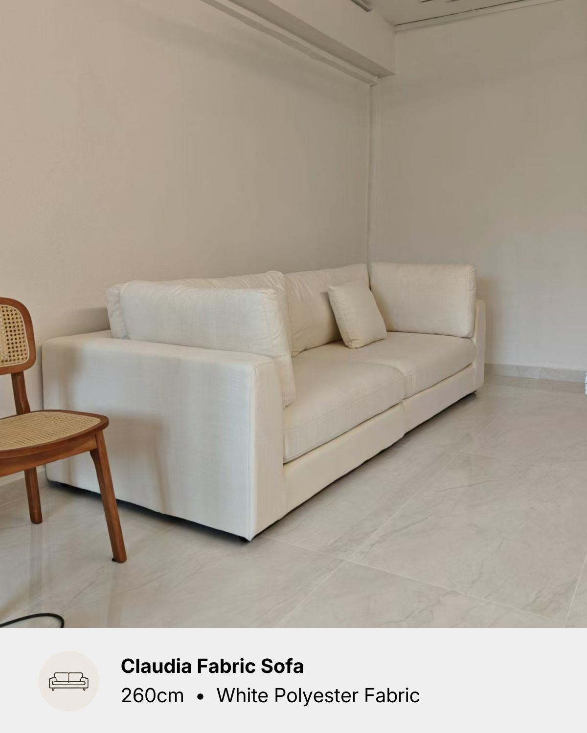 A white Claudia fabric sofa with matching throw pillows, shown in a minimalist room with light-colored walls and tiled flooring. A wooden chair is partially visible on the left.