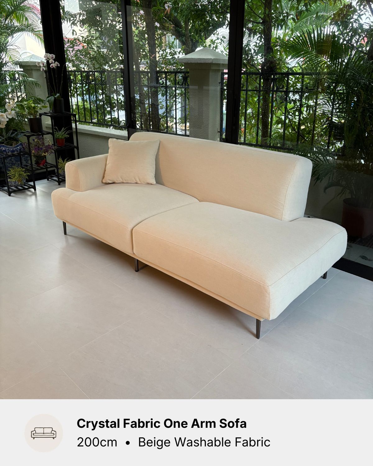 A cream-beige fabric one-arm sofa with a matching back cushion, shown in an indoor showroom setting with large windows and greenery outside. Text at the bottom reads 'Crystal Fabric One Arm Sofa, 200cm • Beige Washable Fabric'.
