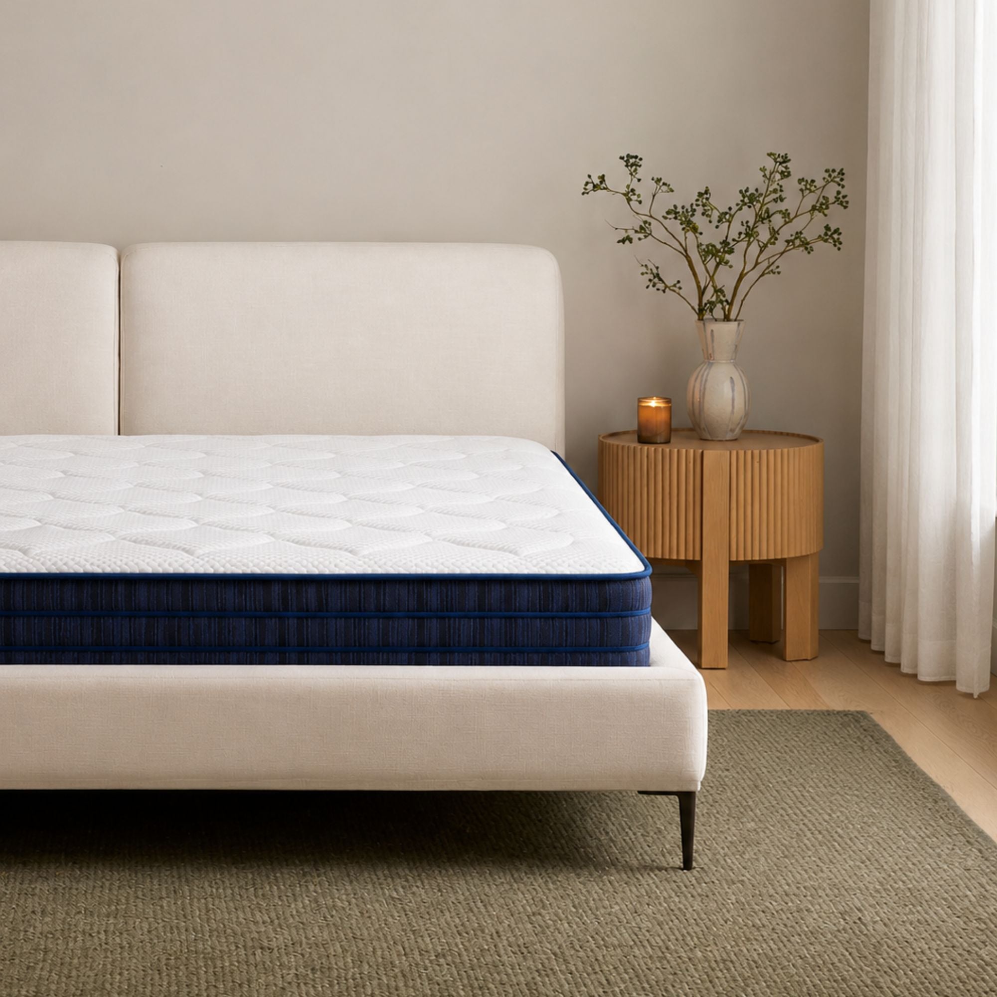 A mattress with a white quilted top and a deep blue side panel, shown on a low upholstered bed in a softly styled bedroom with a beige headboard, wood side table, vase with branches, and a curtain by the window.