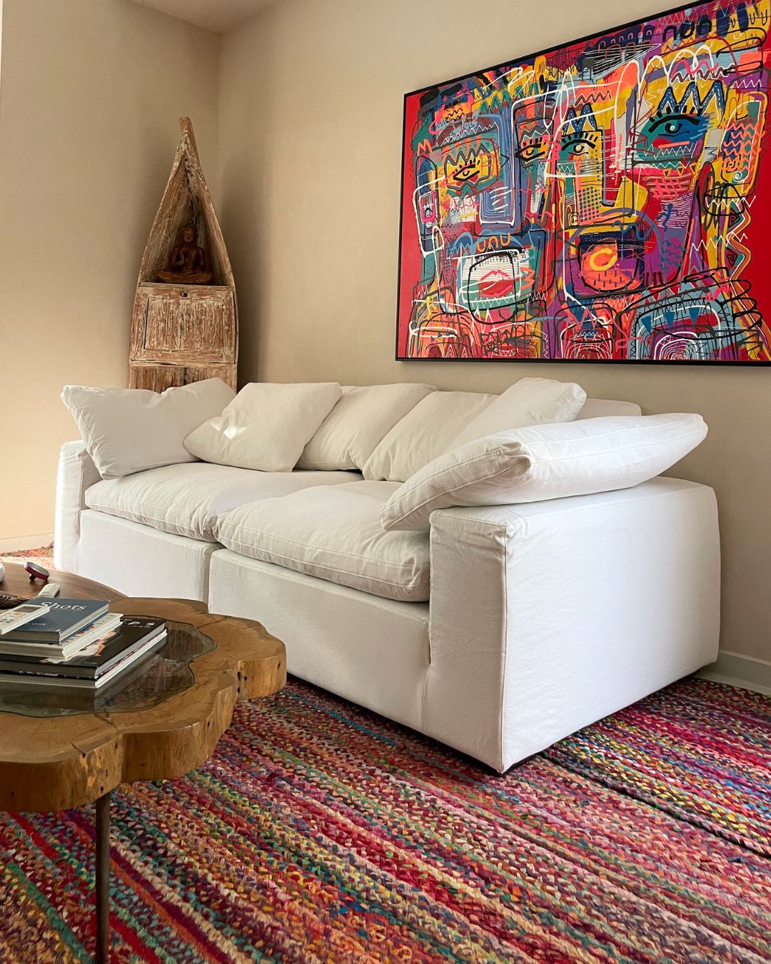 A white upholstered 4 seater sofa in a styled living room, with a colorful abstract wall painting above it, a patterned rug underneath, and a small wooden decorative object in the corner.
