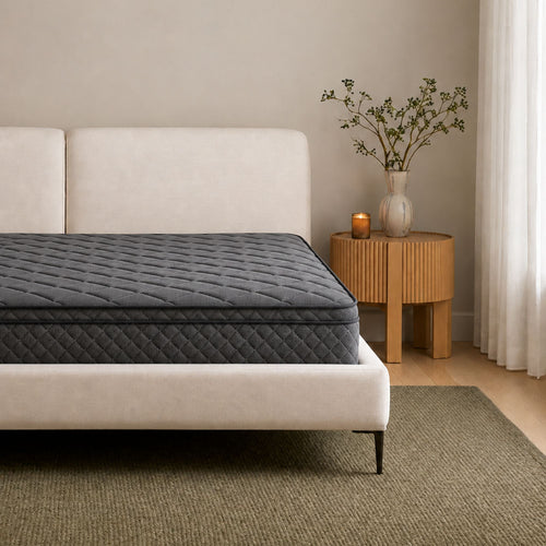 A dark gray quilted mattress on a white upholstered bed frame, styled in a minimalist bedroom with a wooden side table, vase, and curtain.