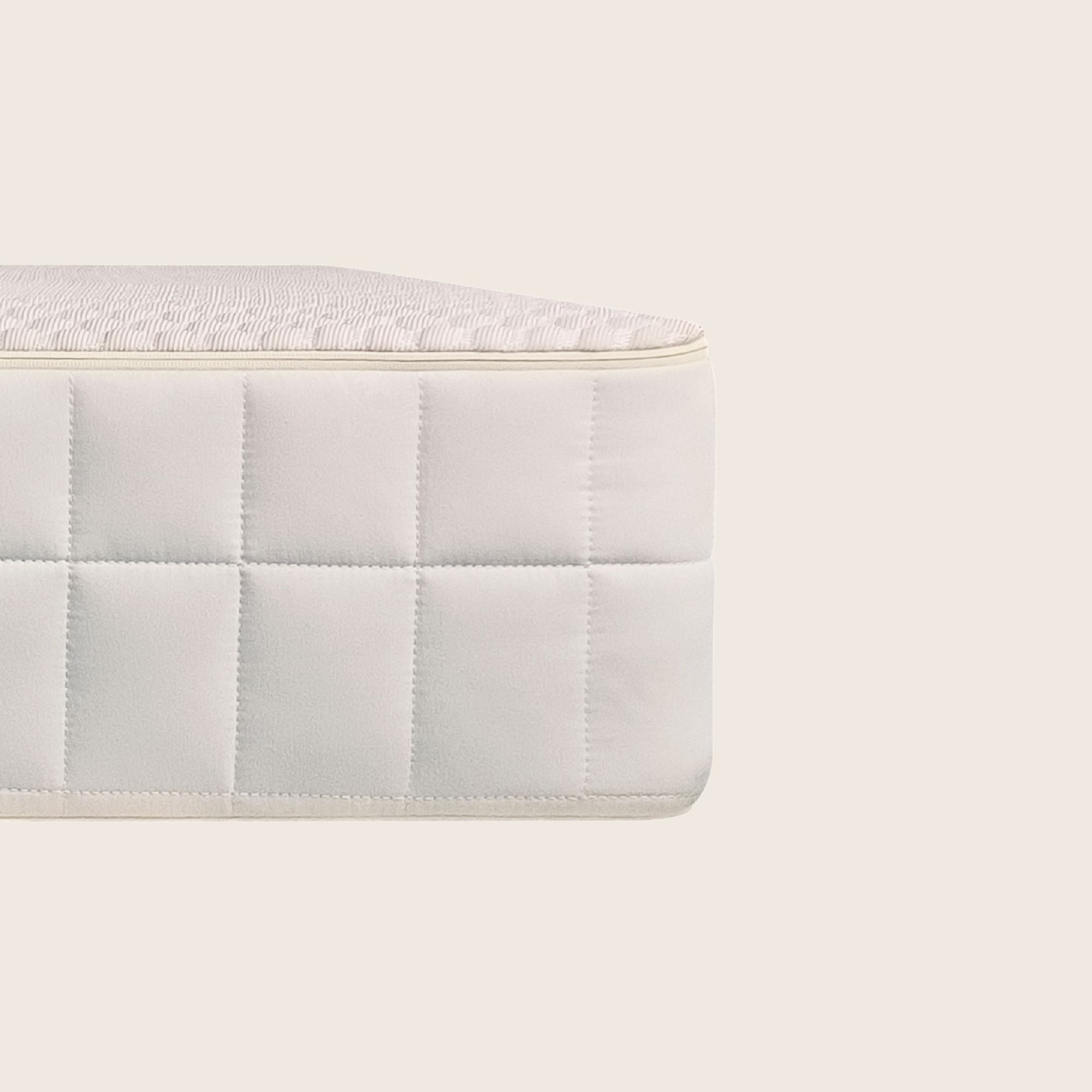A close-up of a white quilted mattress corner with a textured top surface, set against a solid light beige background.