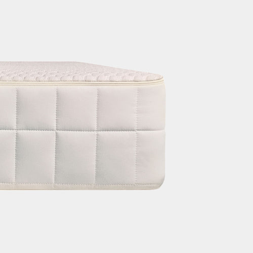 Close-up of a quilted mattress corner showing the side panel and top edge in a light cream color against a plain light gray background.