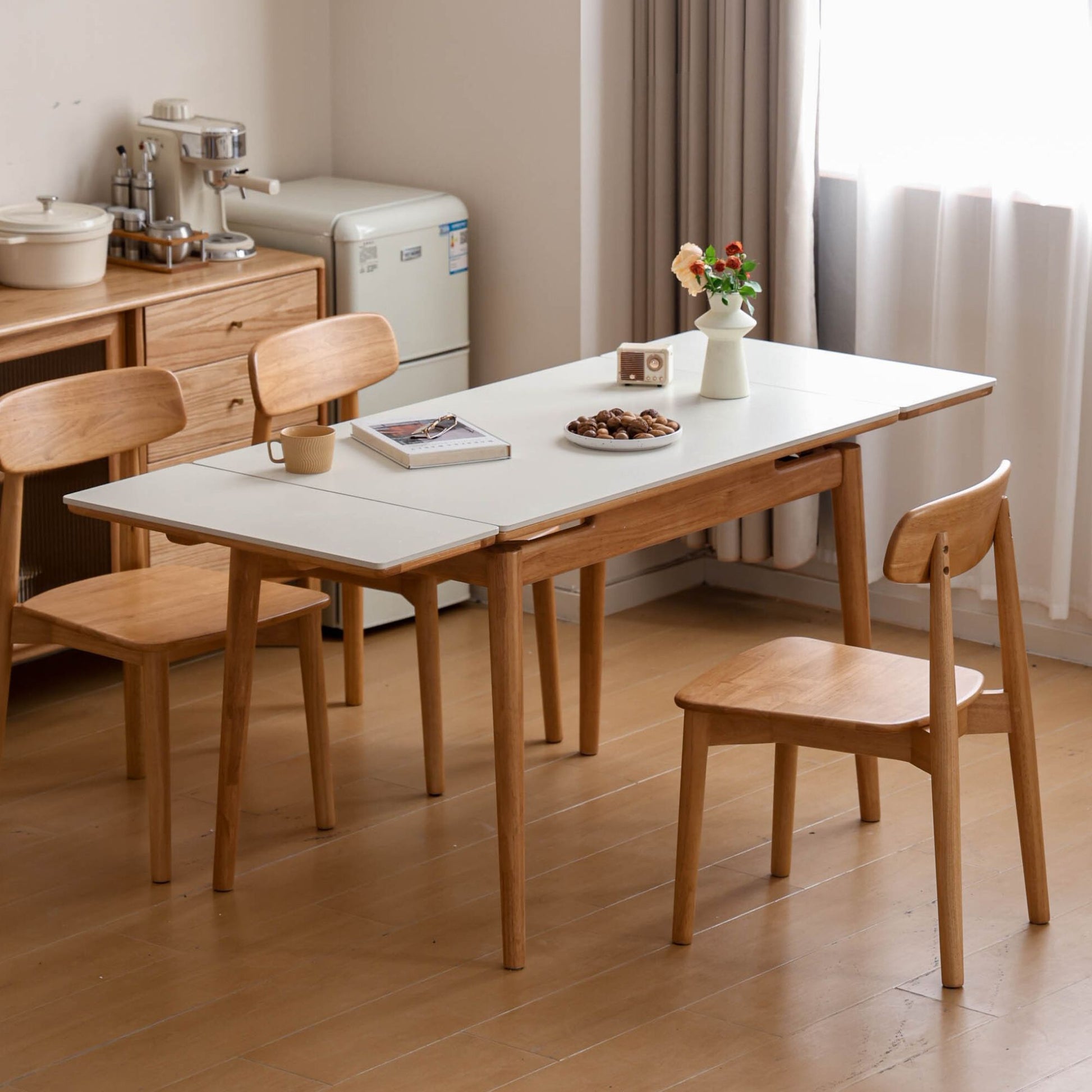 Dining area with a white table and wooden chairs in a room with a refrigerator and cabinets.